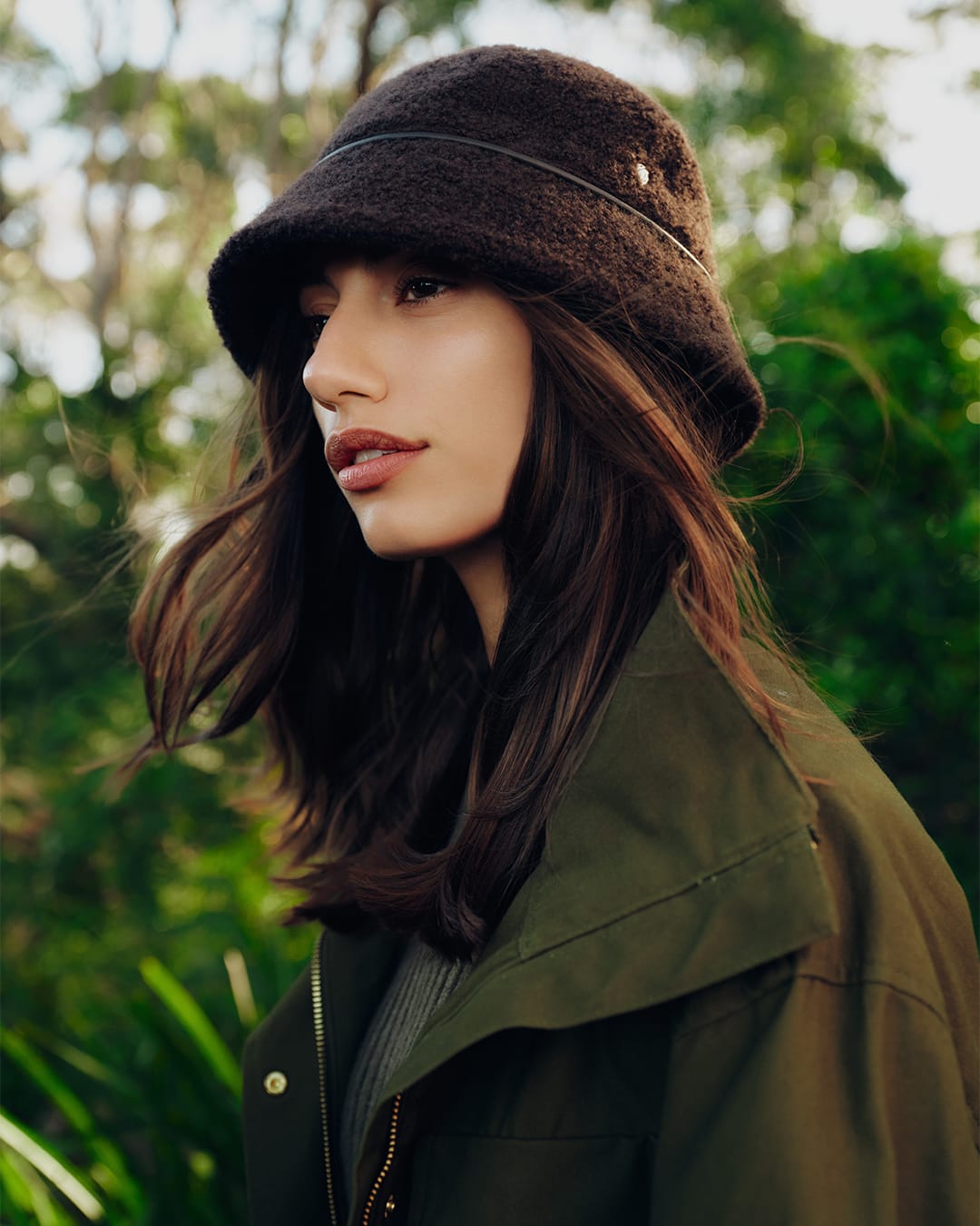 Model wearing a dark brown textured bucket hat and an olive green jacket outdoors in a wooded environment.
