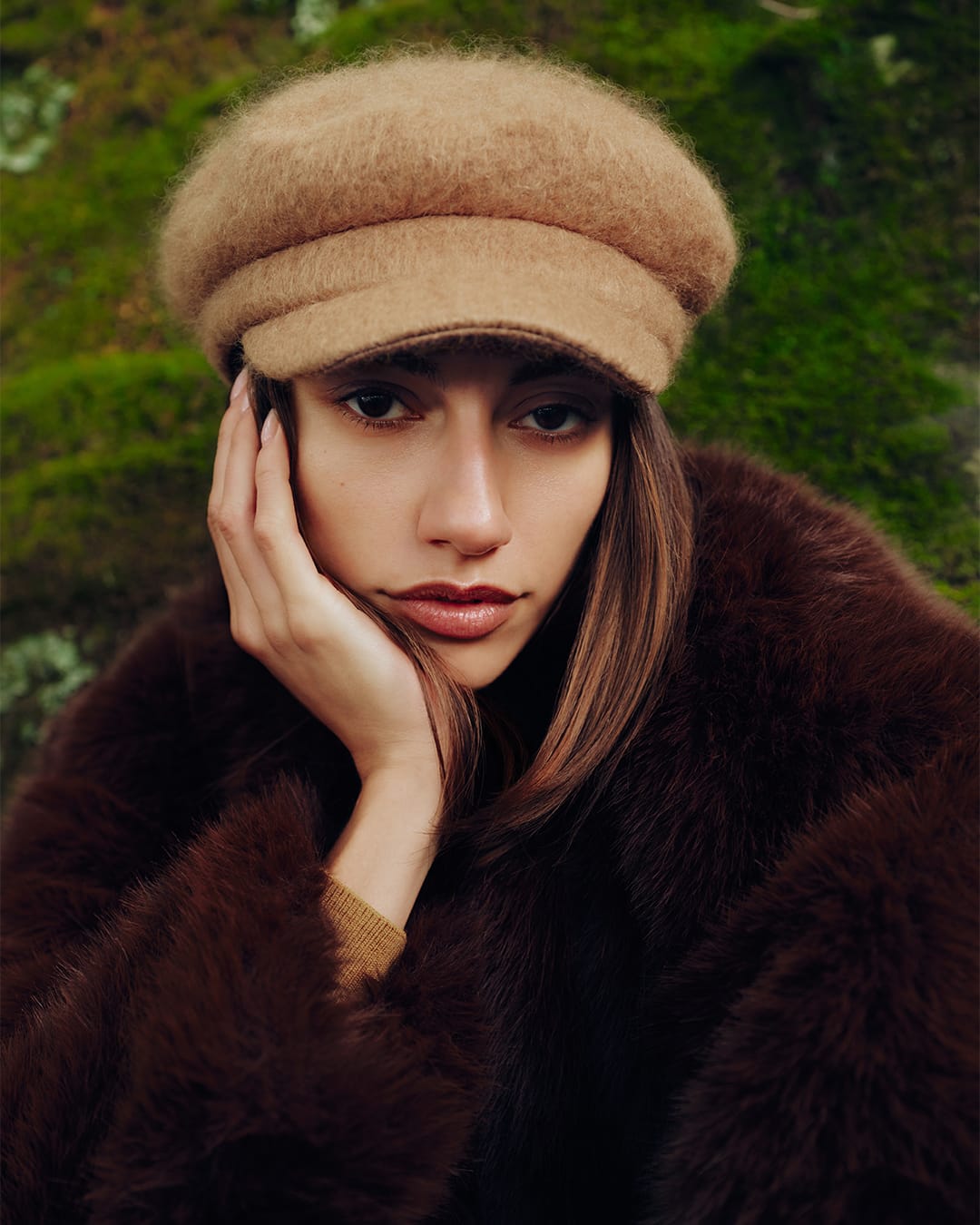 Model wearing a fuzzy camel-colored cap and dark brown textured coat against a mossy green background.