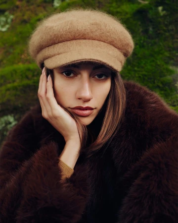 Model wearing a fuzzy camel-colored cap and dark brown textured coat against a mossy green background.