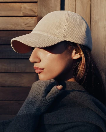 Profile portrait of a woman wearing a beige corduroy baseball cap and a dark grey knit top against a wooden wall background