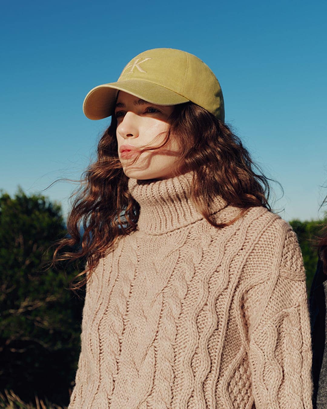 Woman wearing a yellow baseball cap and beige turtleneck sweater against a clear blue sky.