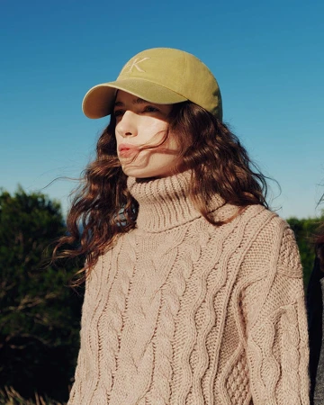 Woman wearing a yellow baseball cap and beige turtleneck sweater against a clear blue sky.
