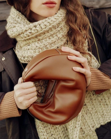 Close-up of a model wearing a thick cream knit scarf holding a brown leather handbag