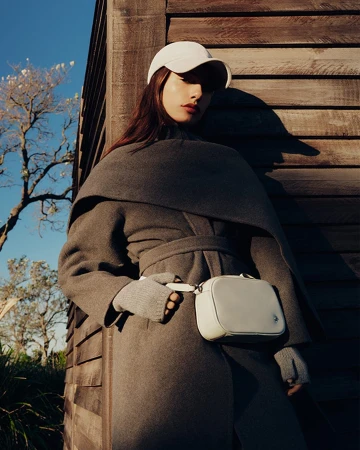Model wearing grey coat, white baseball cap, and crossbody bag leaning against wooden slatted wall outdoors in sunlight