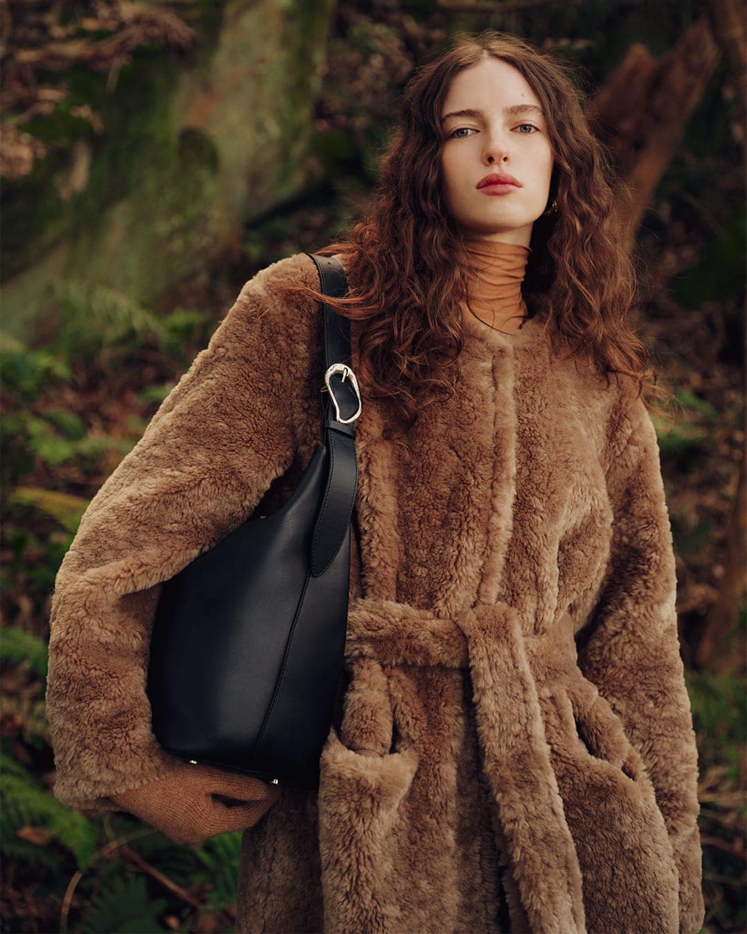 Model with long curly hair wearing a brown fleece coat carrying a black leather shoulder bag in a forest
