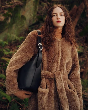 Model with long curly hair wearing a brown fleece coat carrying a black leather shoulder bag in a forest
