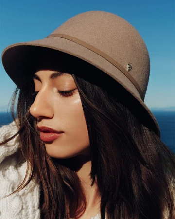 Close-up portrait of a woman wearing a beige felt hat and white knit sweater against a clear blue sky and ocean background