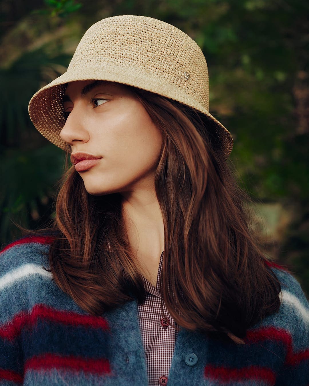 Woman wearing a woven straw hat and a blue and red striped cardigan outdoors against foliage