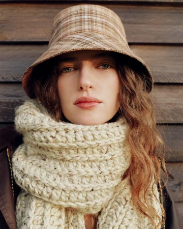 Model wearing a plaid bucket hat and a thick cream knit scarf against dark wooden paneling for Helen Kaminski