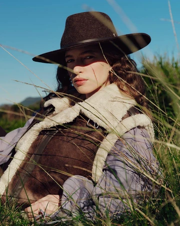 Model wearing a brown fedora hat and shearling vest outdoors in tall grass under a clear blue sky