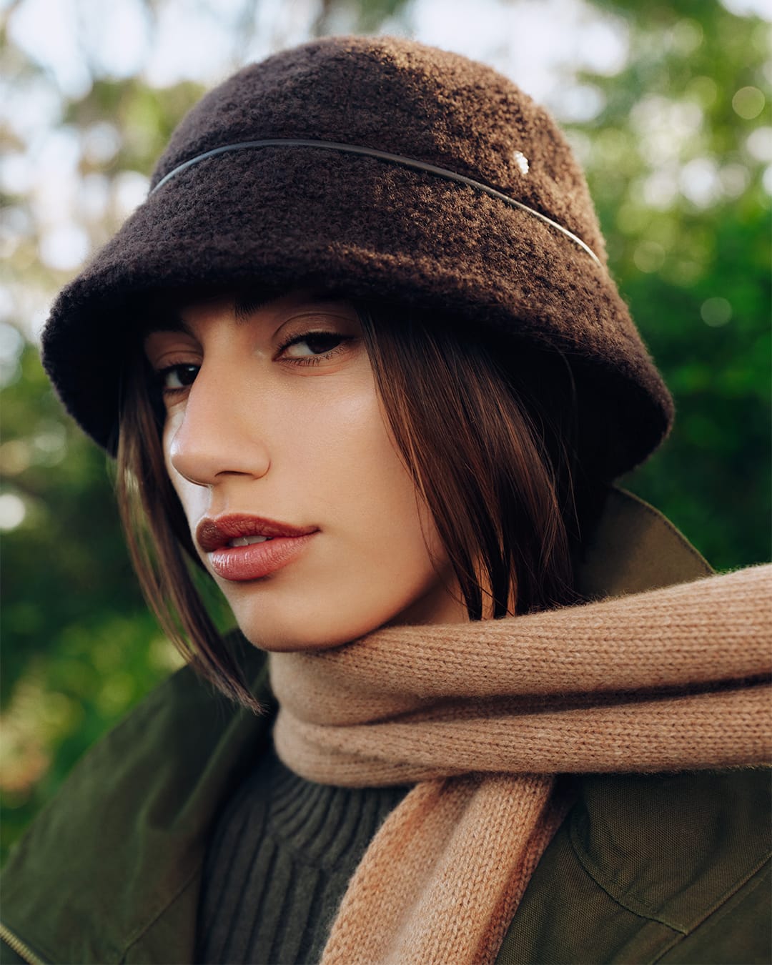 Close-up portrait of a woman wearing a dark brown textured bucket hat and a tan scarf outdoors.