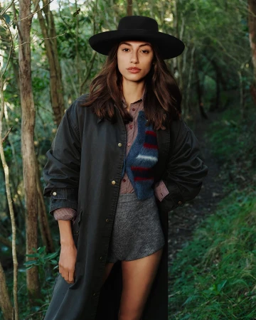 Model wearing a wide-brimmed black hat and dark coat standing outdoors in a lush forest path setting