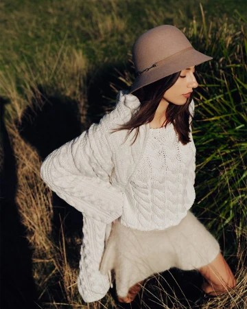 Woman wearing a beige felt hat and white knit cardigan posing outdoors in tall dry grass under natural light.