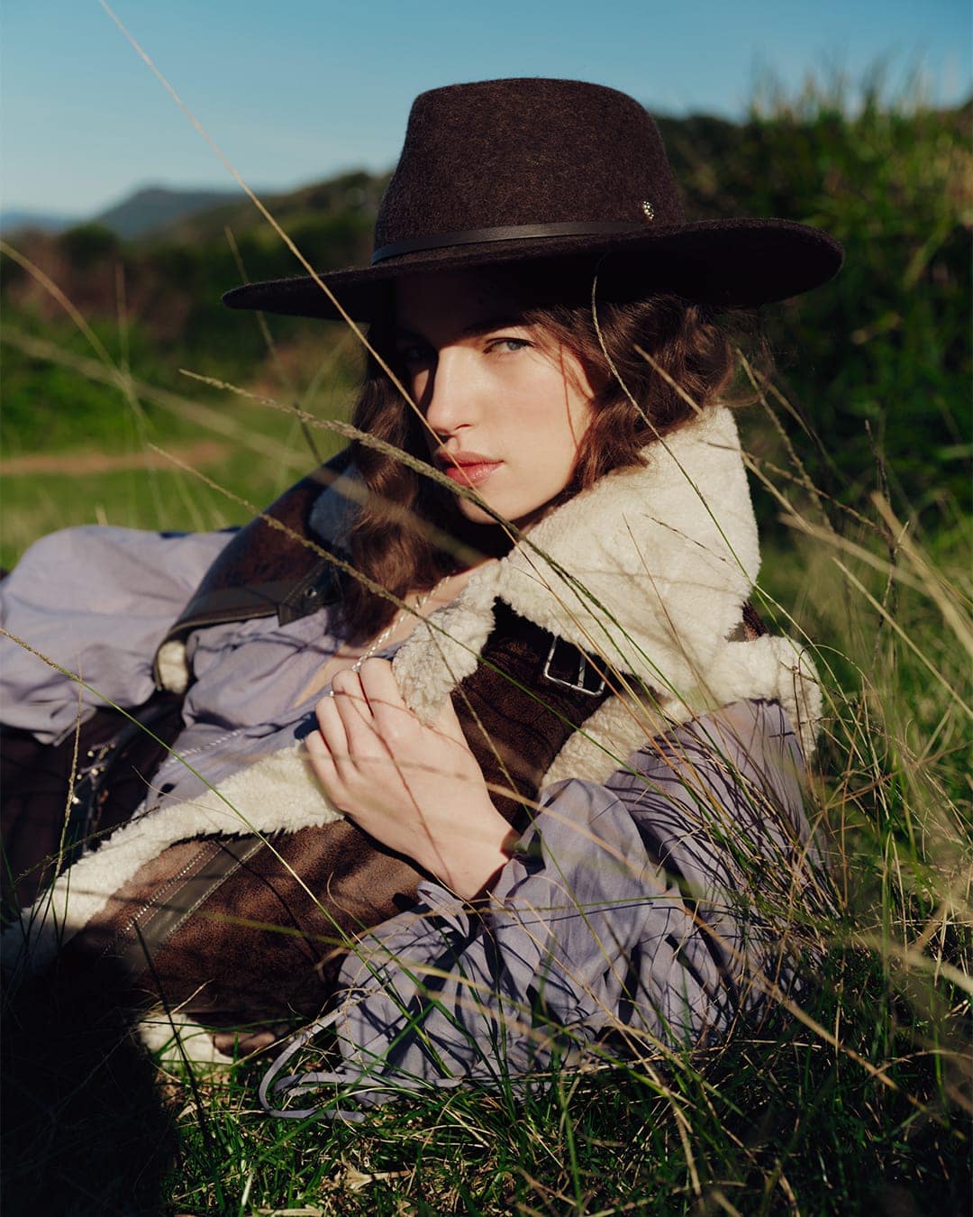 A woman wearing a dark felt fedora and shearling-lined vest lying in tall green grass outdoors under natural sunlight.