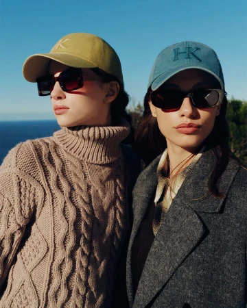 Two women wearing caps and sunglasses outdoors against a clear blue ocean background for Helen Kaminski