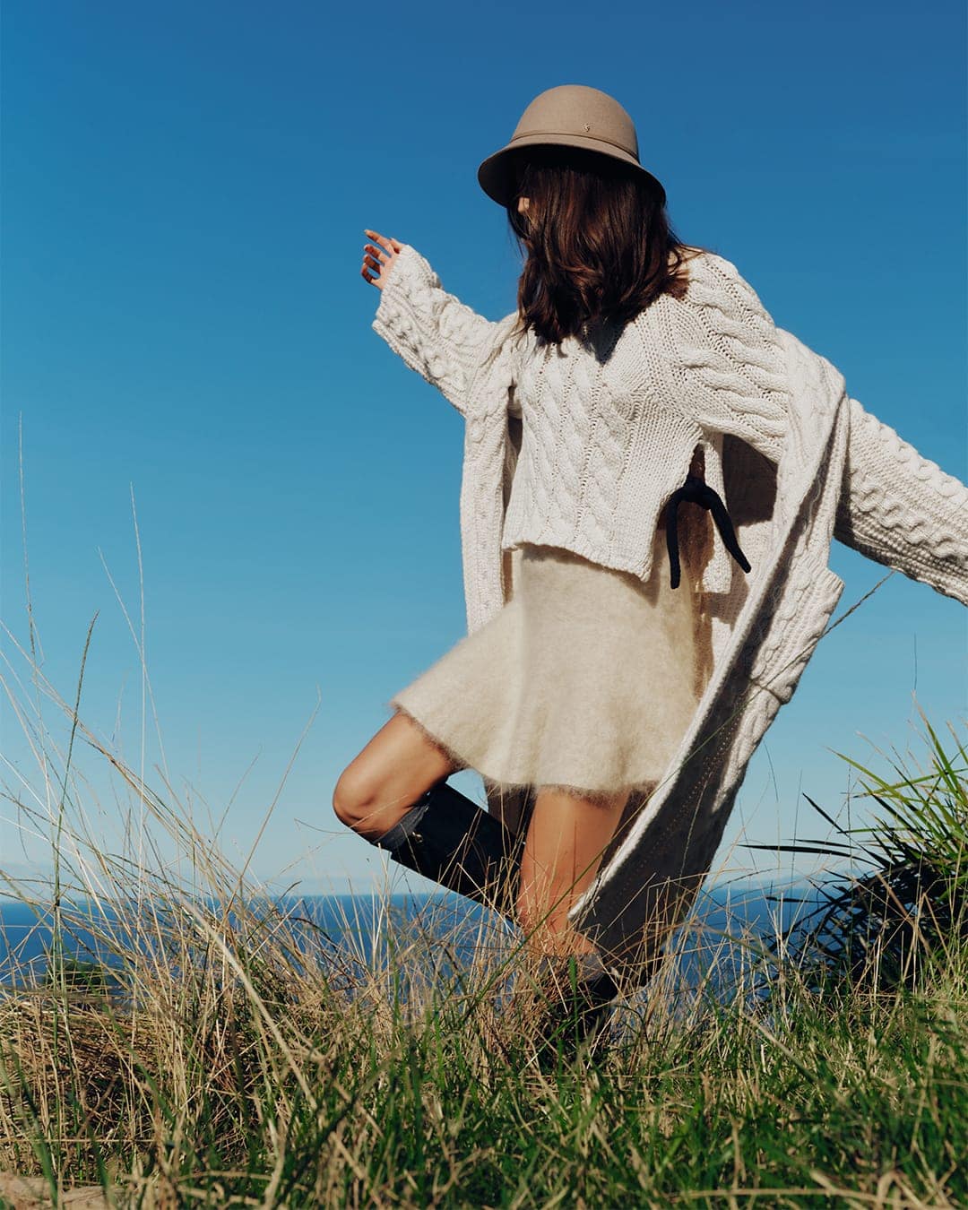 Model wearing a tan hat, white cable-knit sweater, and fuzzy skirt posing outdoors against a clear blue sky