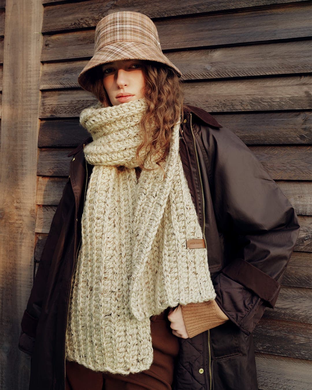 Model wearing a plaid bucket hat and thick knitted scarf standing outdoors against wooden paneling.