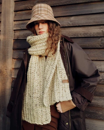 Model wearing a plaid bucket hat and thick knitted scarf standing outdoors against wooden paneling.