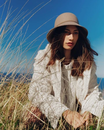 Model wearing a tan felt hat and textured white knit sweater crouches in tall dry grass under a blue sky.