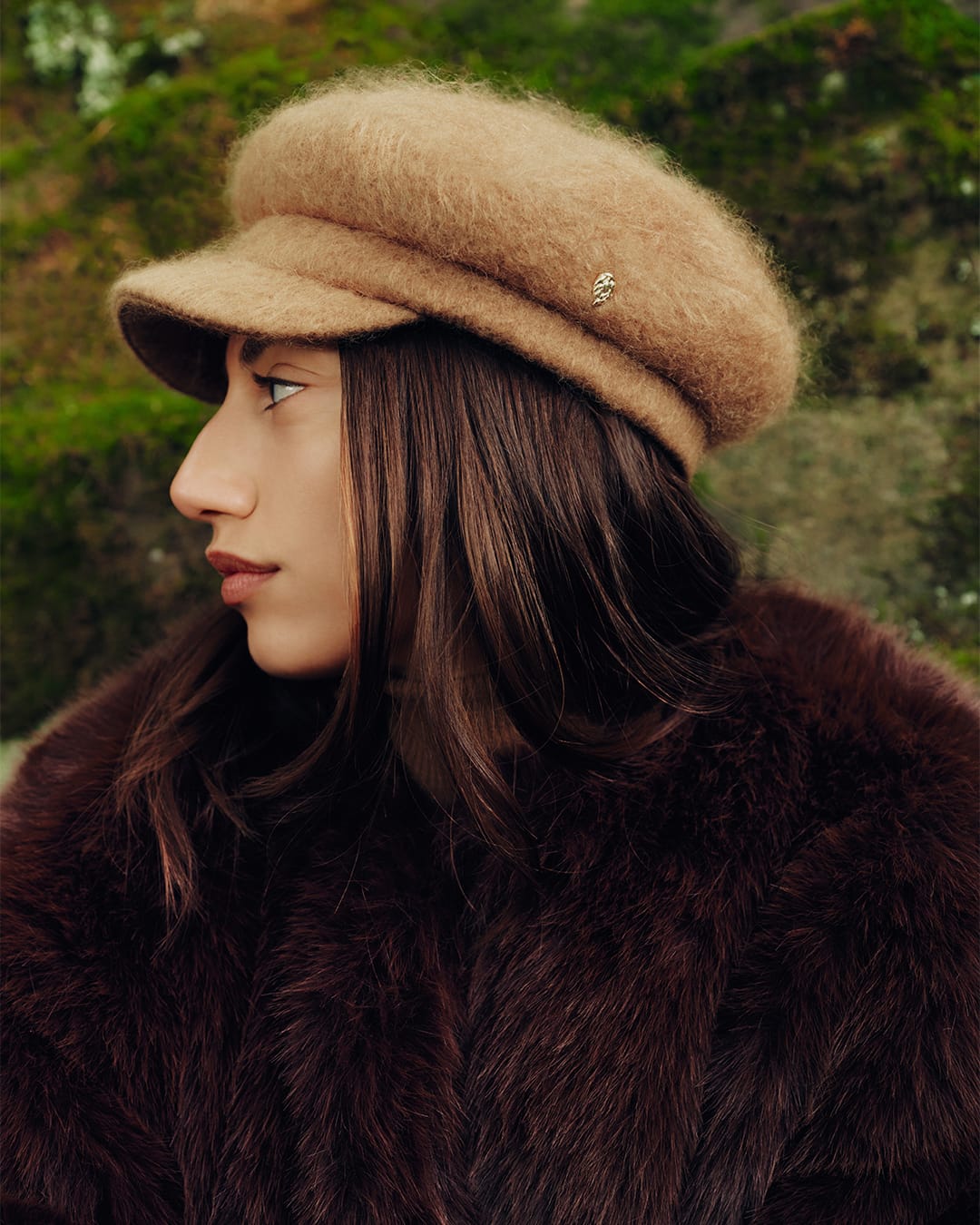 Profile portrait of a woman wearing a fluffy camel cap and a dark furry coat against an out-of-focus natural backdrop.