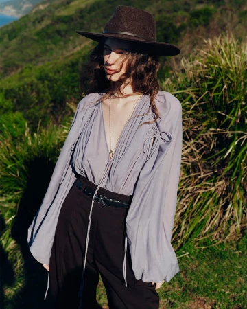 Model wearing a brown fedora hat and a lavender blouse standing outdoors near grassy coastal terrain