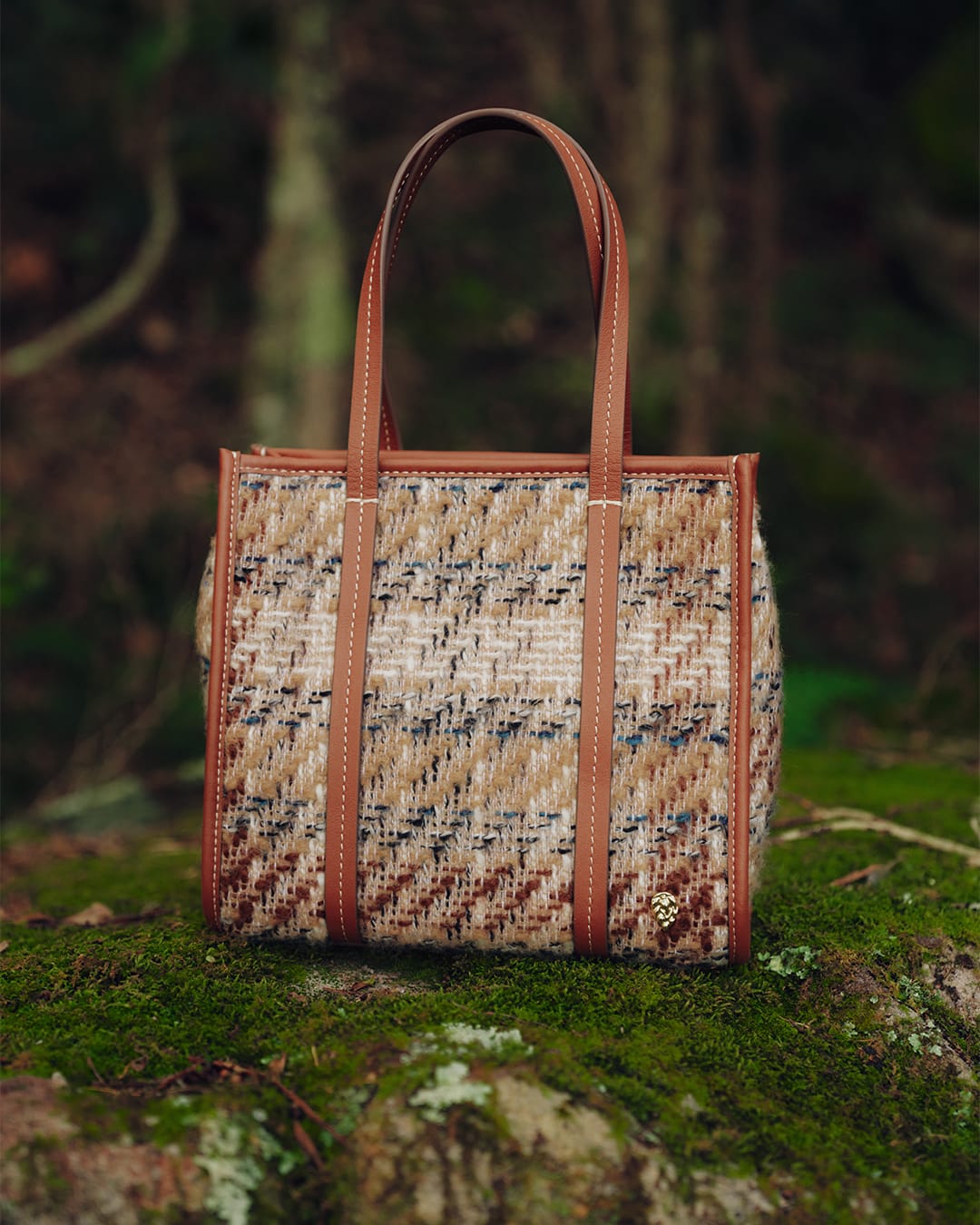 Textured tweed tote bag with brown leather handles resting on a moss-covered rock in a forest setting.
