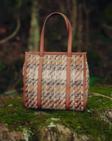 Textured tweed tote bag with brown leather handles resting on a moss-covered rock in a forest setting.