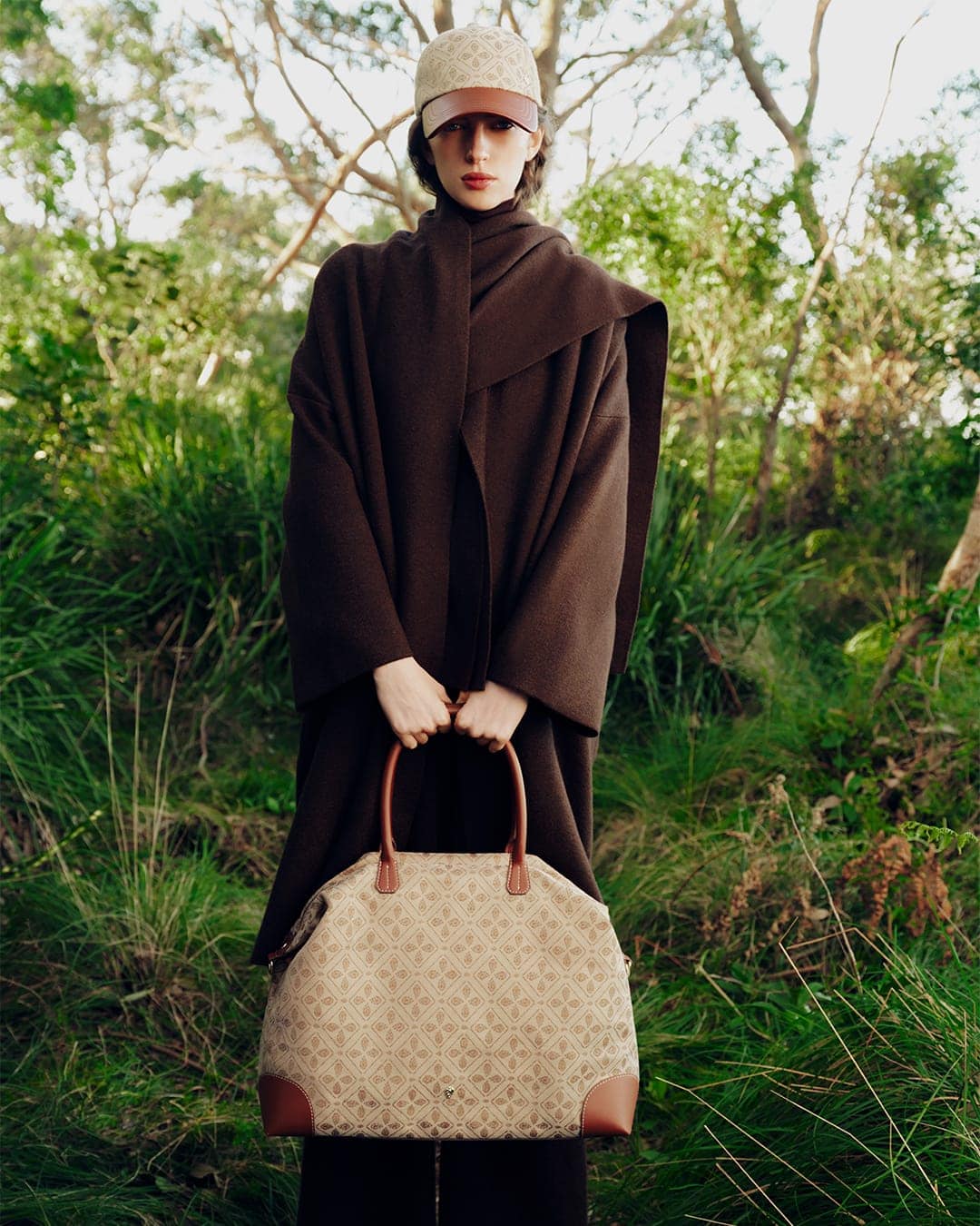 Model in brown wrap coat wearing a patterned cap and holding a large monogram handbag in a wooded forest setting.