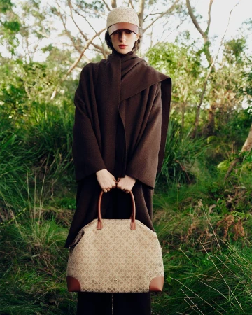Model in brown wrap coat wearing a patterned cap and holding a large monogram handbag in a wooded forest setting.