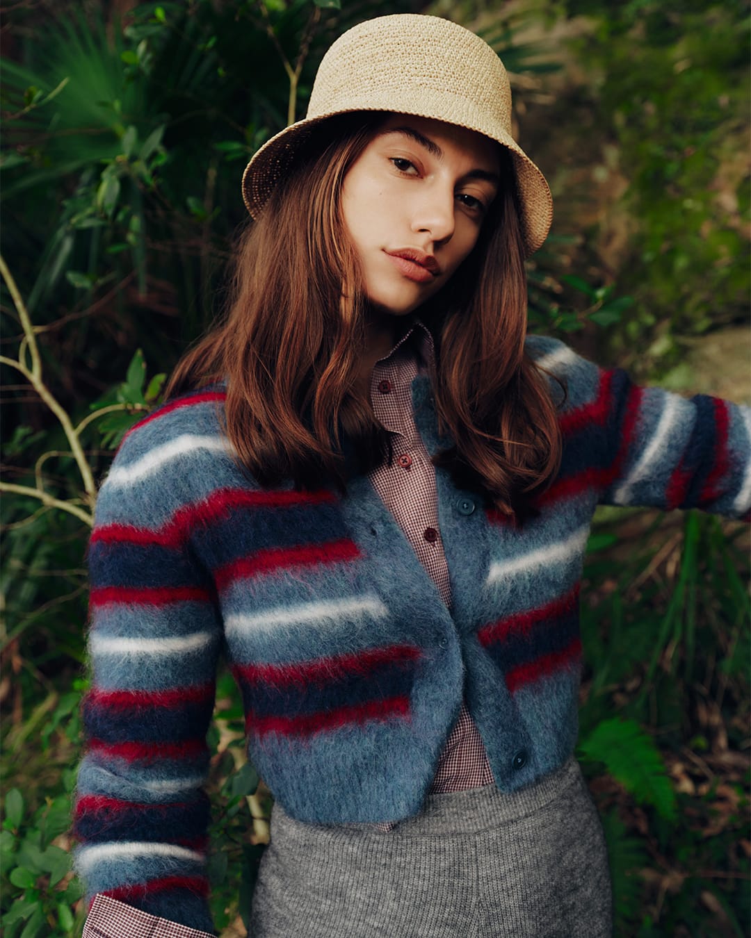 Model wearing a straw bucket hat and a blue striped cardigan against natural foliage in a campaign portrait