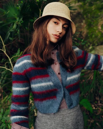 Model wearing a straw bucket hat and a blue striped cardigan against natural foliage in a campaign portrait