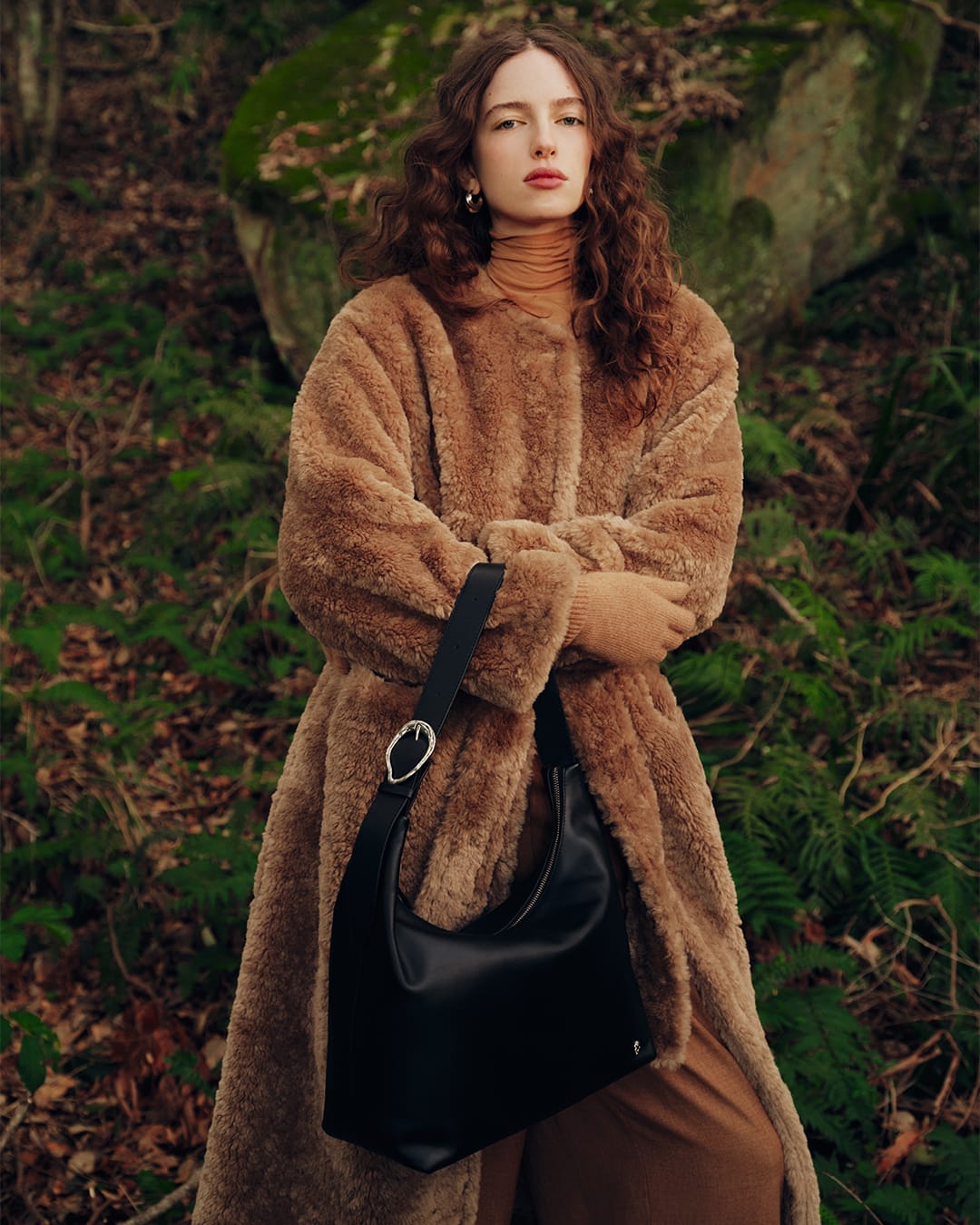 Model with long curly brown hair wearing a fluffy brown coat holding a black handbag in a lush forest setting.