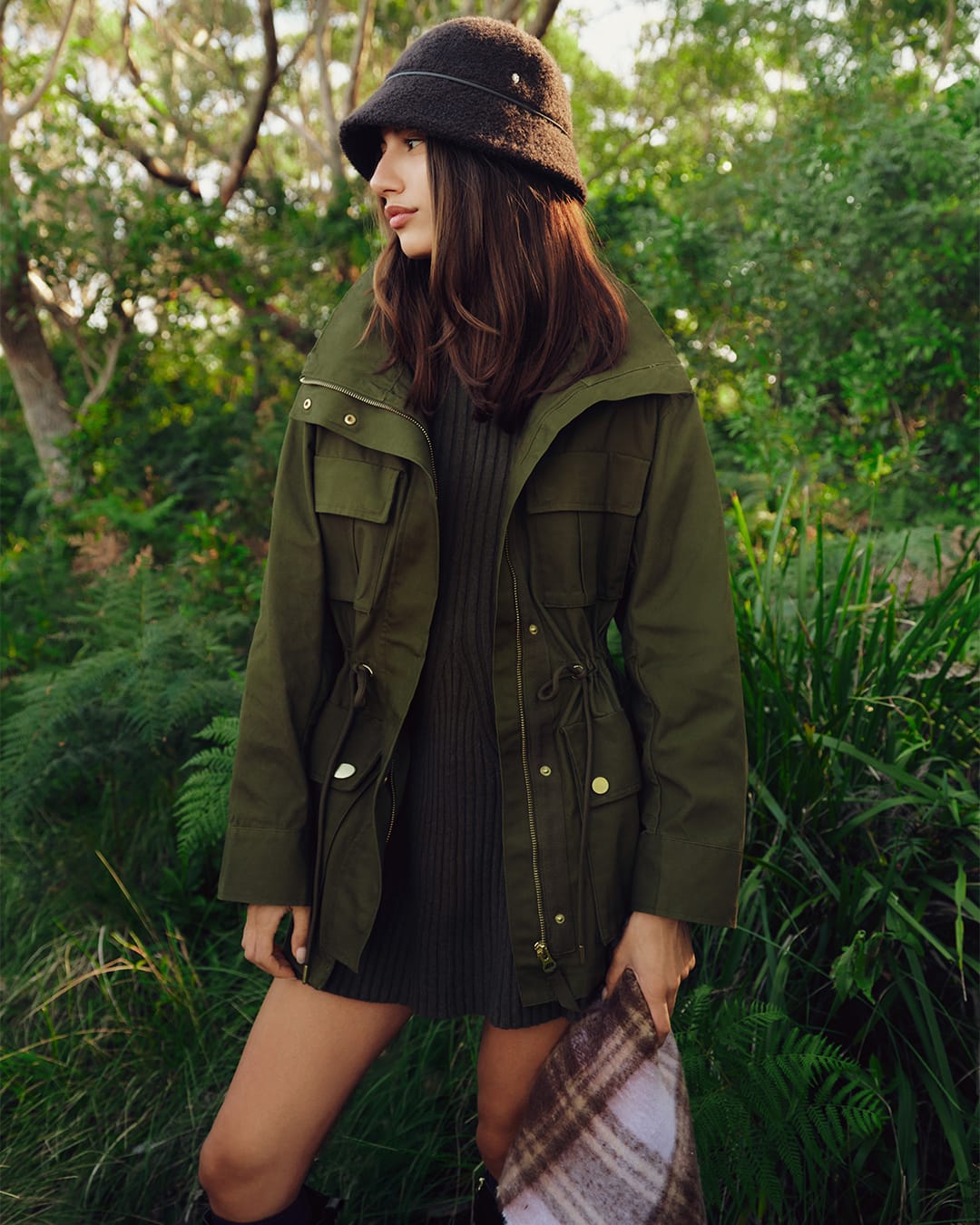 Woman wearing an olive green jacket and dark bucket hat standing outdoors amidst lush green ferns and foliage.