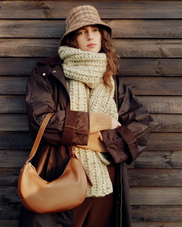 Model wearing a plaid bucket hat, chunky knit scarf, and dark quilted coat holding a tan leather shoulder bag against wood paneling.