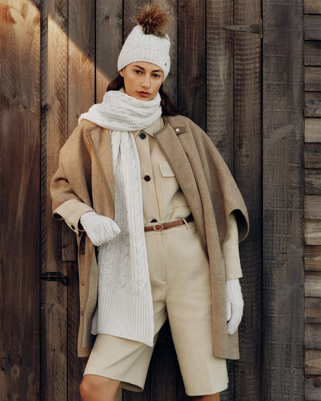 Model wearing a knit beanie, scarf, and beige cape coat standing against a wooden vertical slat wall.