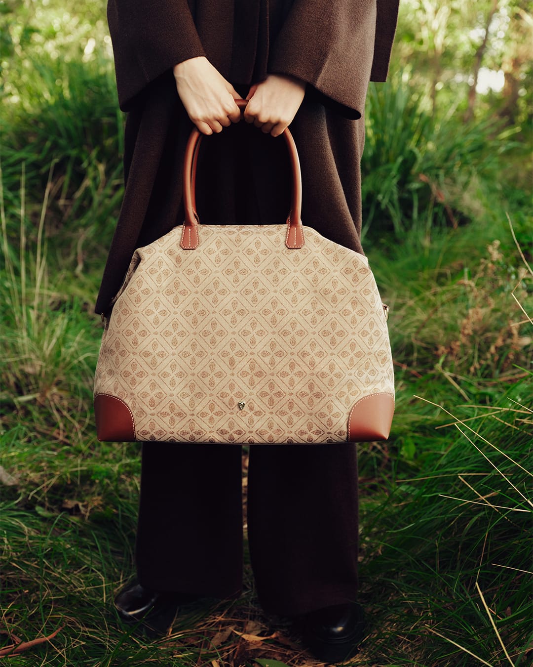 Person holding a patterned canvas handbag with leather handles standing outdoors in a grassy environment