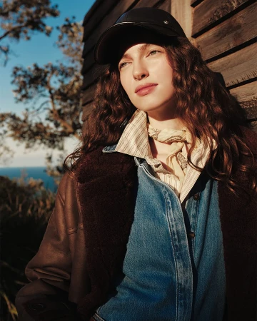 Woman wearing a leather cap, striped shirt, and brown shearling jacket outdoors against a wooden wall and ocean view.