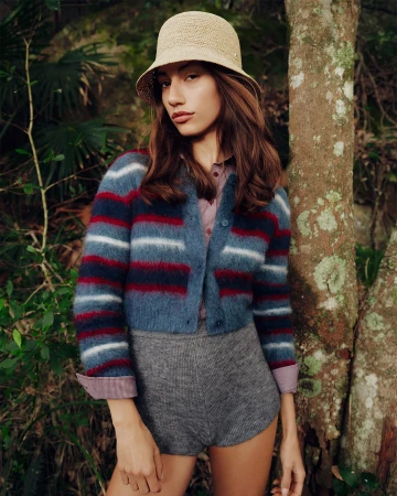 Model wearing a striped cardigan, grey shorts, and a straw bucket hat standing next to a tree in a forest setting.
