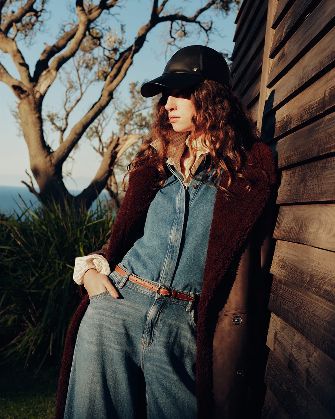Model wearing a black cap, denim shirt, and burgundy coat leaning against a wooden wall outdoors at sunset.