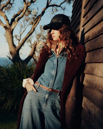 Model wearing a black cap, denim shirt, and burgundy coat leaning against a wooden wall outdoors at sunset.