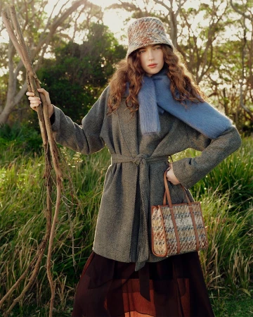 Model wearing grey belted coat, blue scarf, and patterned bucket hat holding sticks outdoors in a grassy field