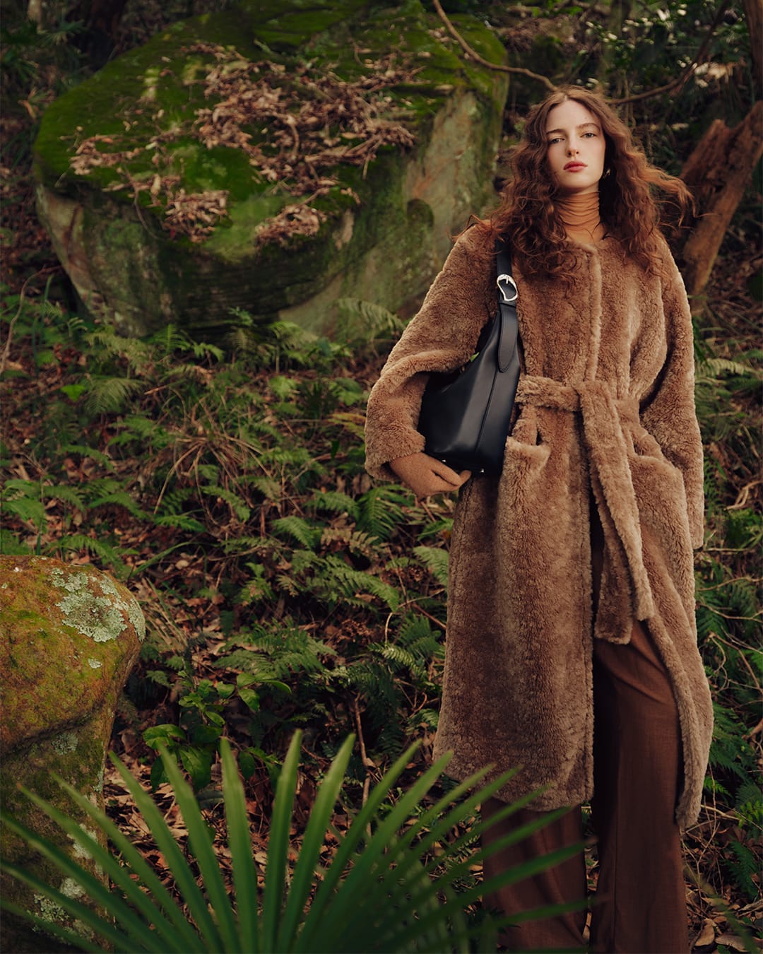 Model wearing a brown textured coat and carrying a black shoulder bag in a forest setting with large mossy rocks.
