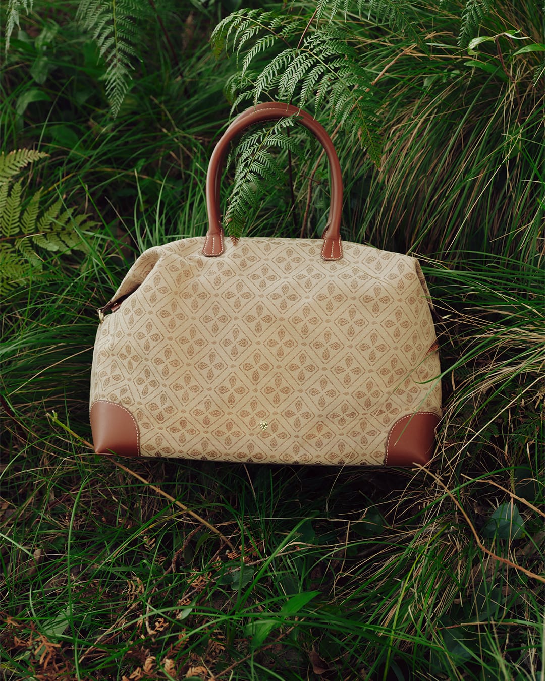 Patterned canvas weekender bag with brown leather handles nestled in lush green grass and ferns.