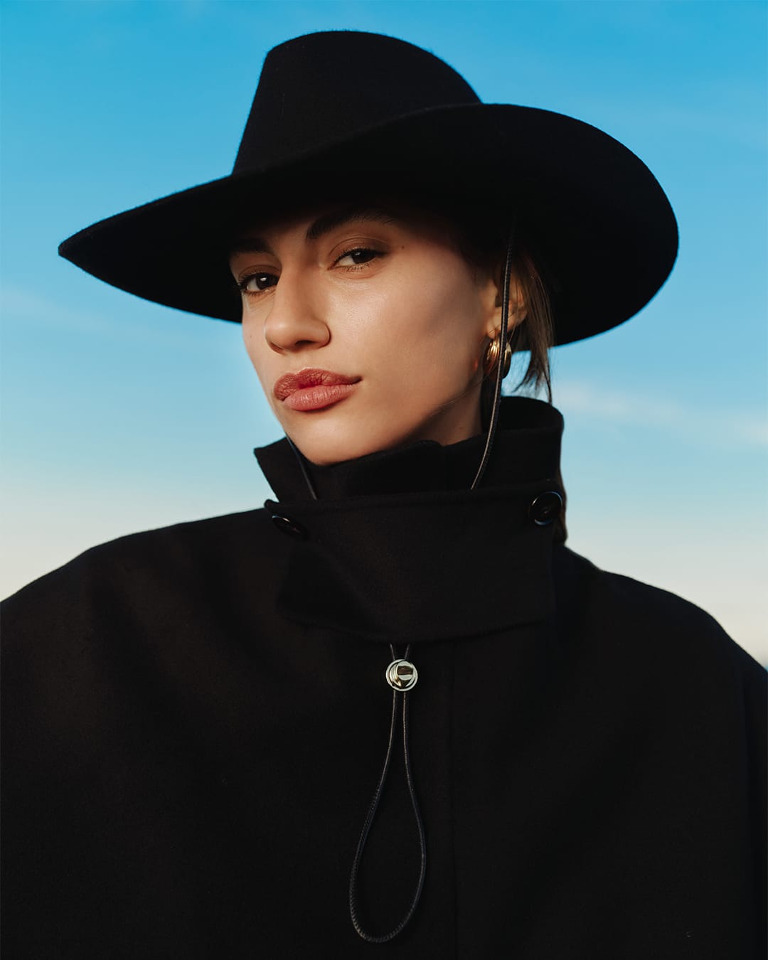 Close-up portrait of a woman wearing a black wide-brimmed hat and black coat against a bright blue sky.