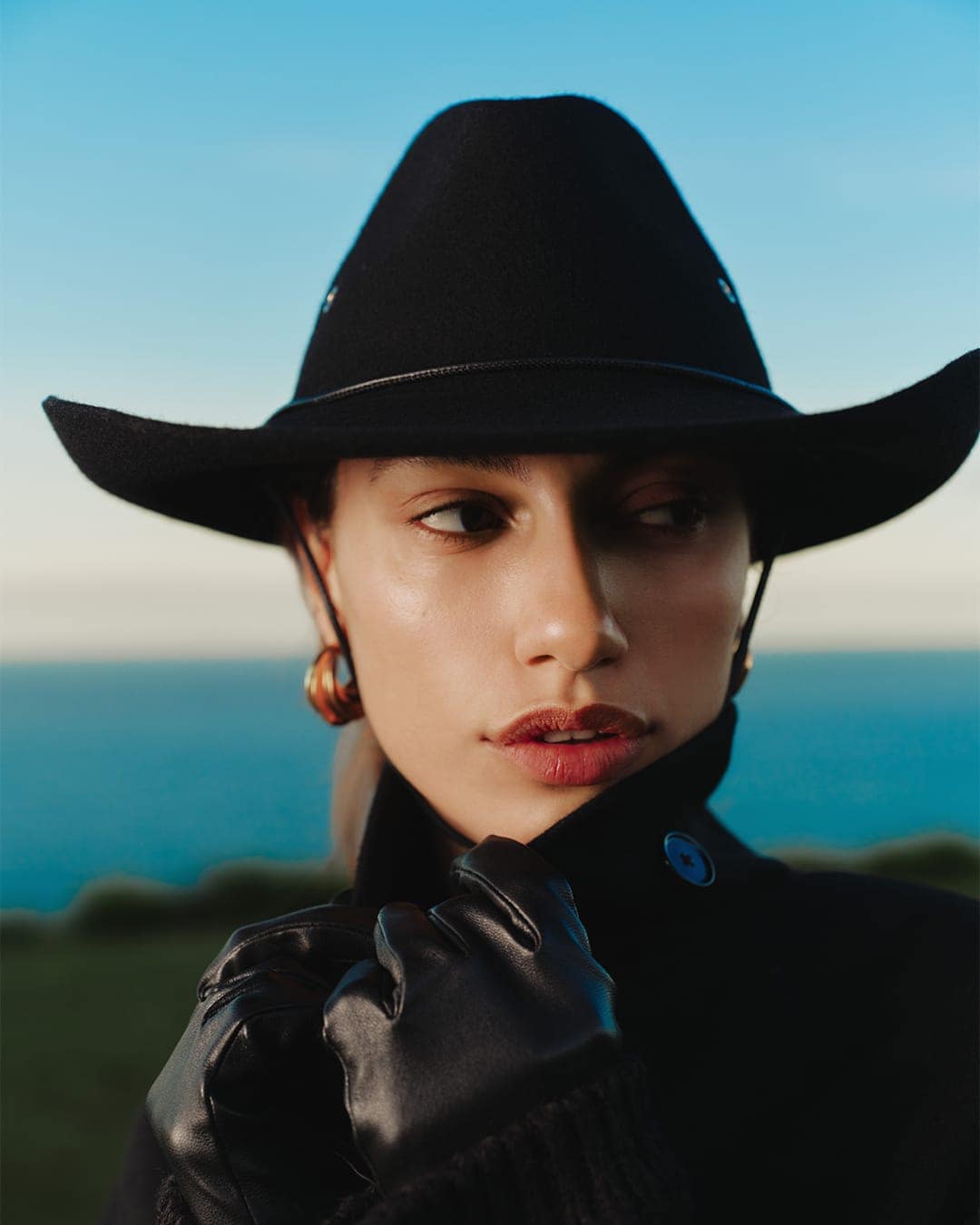 Model wearing a black cowboy hat and leather gloves against an ocean background for Helen Kaminski