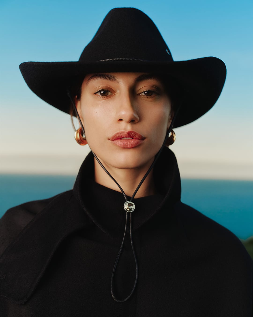 Close-up portrait of a woman wearing a black wide-brim hat and a black coat against an out-of-focus horizon.