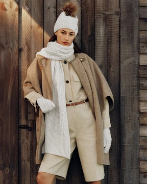 Model wearing a knit beanie, scarf, and beige cape coat standing against a wooden vertical slat wall.