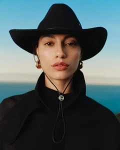 Close-up portrait of a woman wearing a black wide-brim hat and a black coat against an out-of-focus horizon.