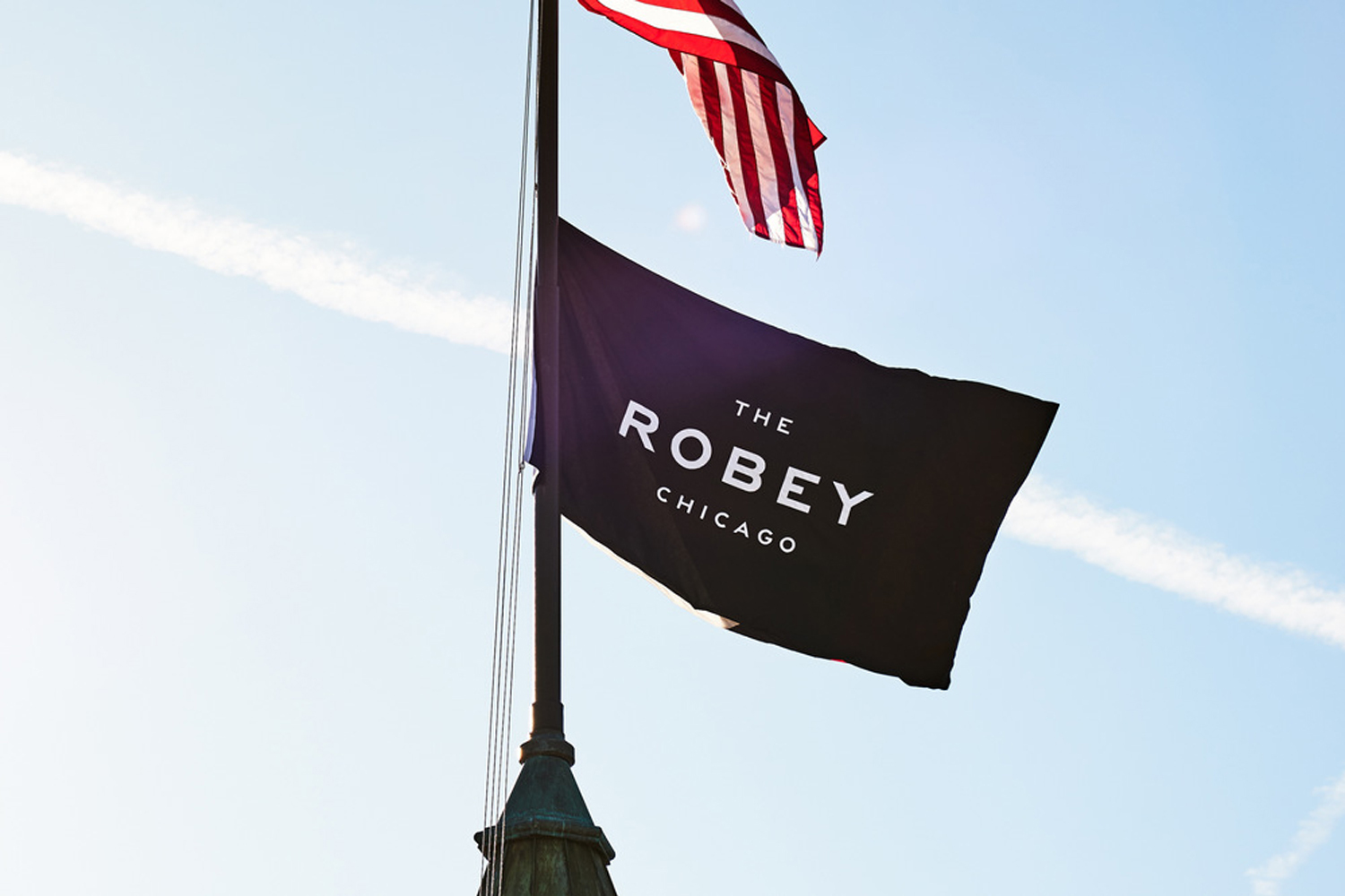 Black flag with white typography reading The Robey Chicago flying on a flagpole beneath an American flag against a clear blue sky