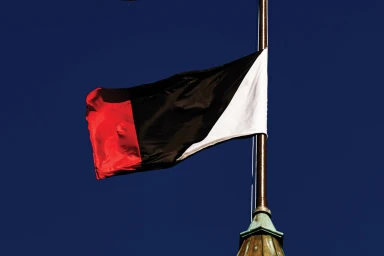 Flag with red, black, and white vertical sections flying on a pole against a deep blue sky.